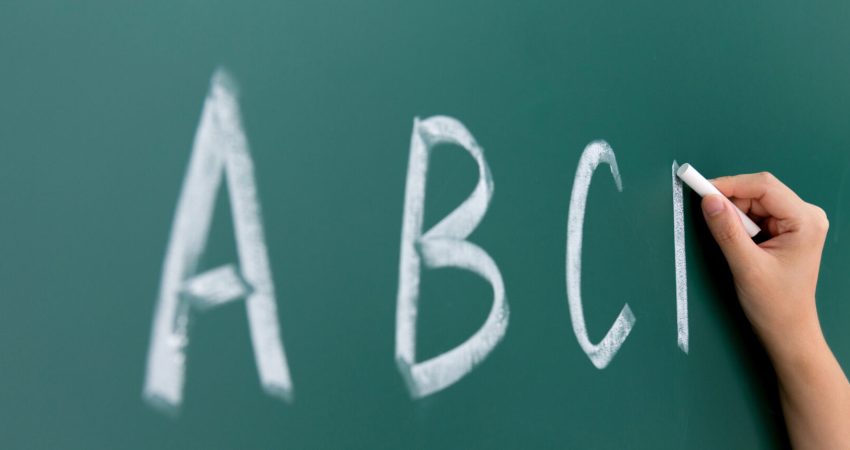 A picture of a hand holding a piece of chalk, writing "A B C" on a chalk board