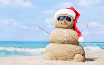 A picture of a snowman made out of sand wearing a Santa hat on a beach with the ocean in the background