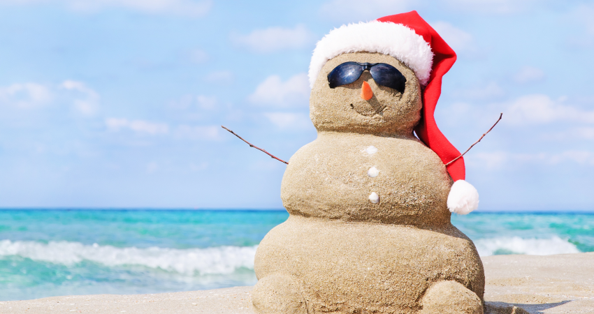 A picture of a snowman made out of sand wearing a Santa hat on a beach with the ocean in the background