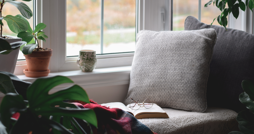 A cozy, relaxing reading area, featuring a cushy chair with a book and some glasses in the middle of it, next to a window,