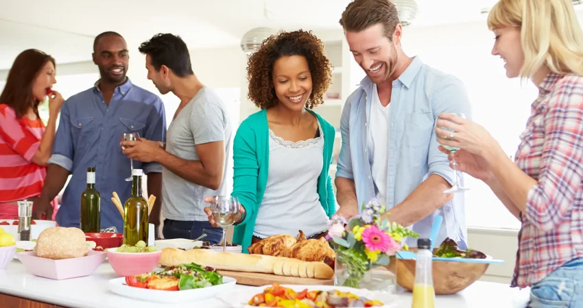 Diverse group of happy friends gathering around a kitchen counter filled with food, roasted chicken, bread, and wine during a dinner party.