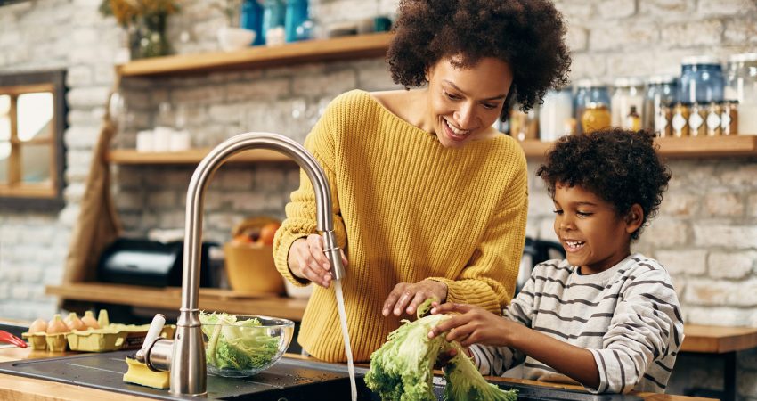 Happy mother and son cleaning vegetables under kitchen sink.