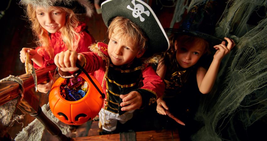 Happy children in carnival costumes trick or treating with pumpkin basket in the old wooden house. Halloween party.