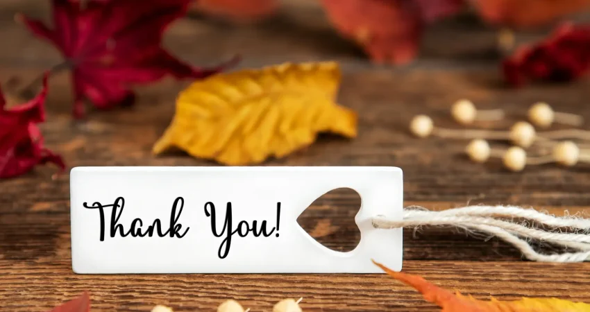 White Thank You tag with a heart cutout on a rustic wooden table surrounded by autumn leaves