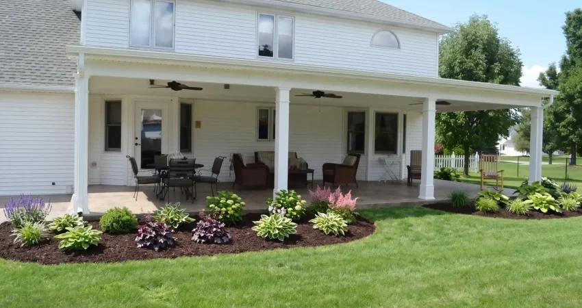 Large covered patio with white columns, outdoor furniture, ceiling fans, and a neatly landscaped garden bed bordering a lush green lawn.